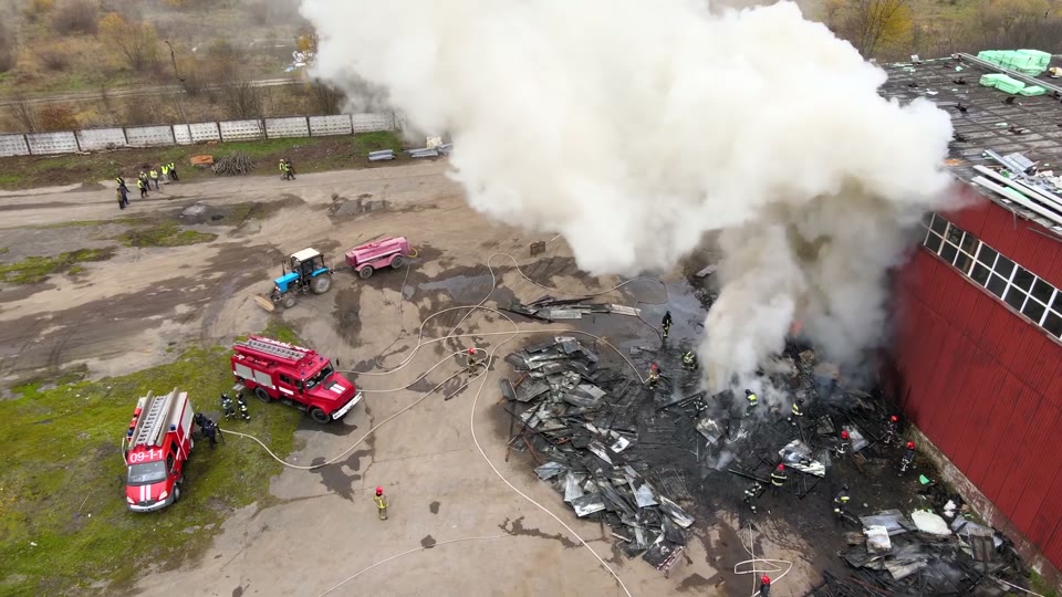 Drone view of an industrial fire response: firefighters on a smouldering debris pile beside a red building, fire engines and a water tanker